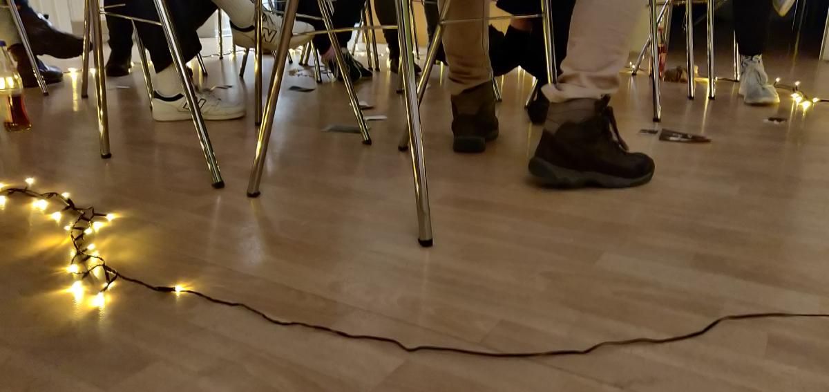 Ein hellbrauner Fußboden auf den metall Stühle stehen, wir sehen verschiedene Füße von Menschen die auf den Stühlen sitzen. Um die Stuhlgruppe herum liegt eine Lichterkette auf dem Fußboden.