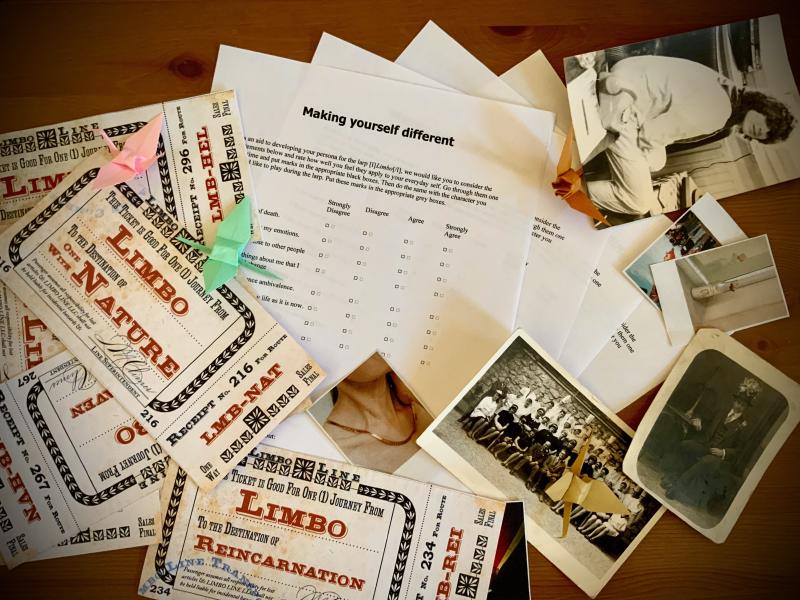 Auf einem Tisch liegen die Spielmaterielen für Limbo. Links ein stapel Fahrkarten auf denen verschiedene Ziele wie “One with Nature” oder “Reincarnation” steht. In der mitte liegen die Charakterblätter die minimalistisch schwarz weiß gehalten sind und einige Multiplechoice Fragen enthalten. Rechts im Bild liegen alte Fotos von Personen, Landschaften und Gegenständen. Darüber sind vier origami Kraniche verteilt in rosa, mintgrün so wie braun.