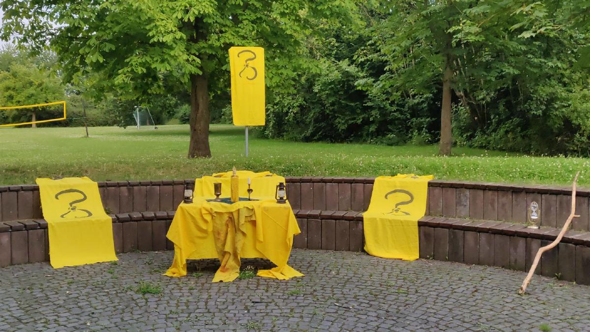 Eine Platz auf einer Wiese mit Sitzbänken im Boden. In der Mitte ist ein Tisch überdeckt mit einem gelben Tuch, darauf steht eine gelbe Kerze, zwei Öllampen, ein Kelch und eine kleine Kerze. Neben dem Tisch, über den Bänken, liegen links und rechts zwei gelbe Tücher mit einem schwarzen Zeichen, drei Halbkreise, die um einen Punkt in der Mitte stehen. Ein drittes Tuch ist weiter hinten als Flagge aufgestellt. Rechts lehnt ein knochiger Stock und eine weitere Laterne.